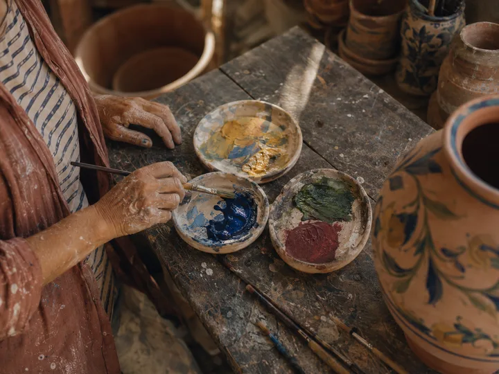 Top-down view of paint palettes and a half-painted terracotta vase on Elena's workbench.