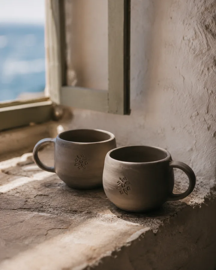Two finished stamped mugs on a Sifnos windowsill, Aegean sea behind.
